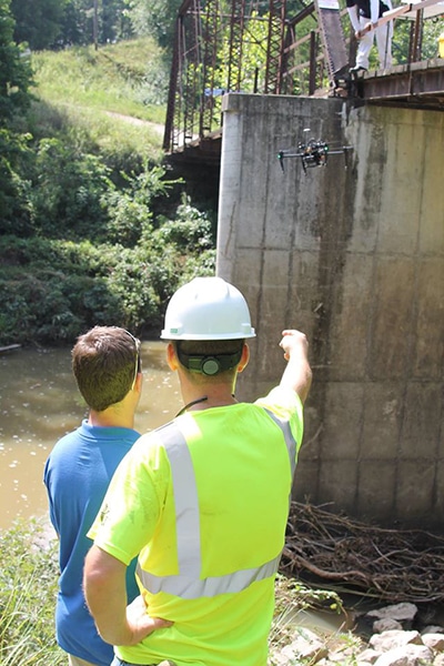 Two men stand in the foreground while piloting a drone near a bridge for inspection